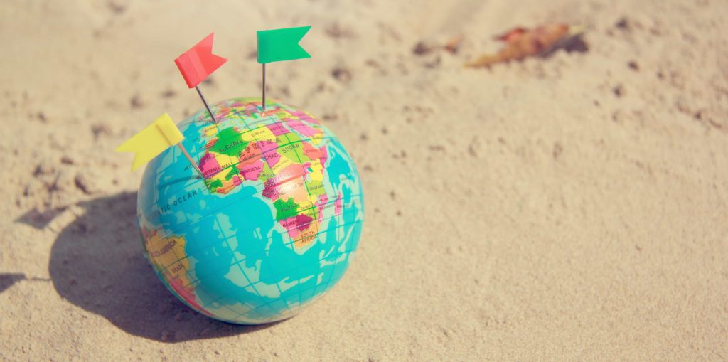 A small globe with colorful pin markers is placed on sandy ground. The globe displays continents and countries with distinct colors, and the pins are red, green, and yellow, inserted into different locations on the globe.