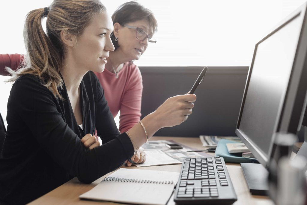 Two women are focused on a computer screen. One, in a black sweater, is pointing at the screen while holding a pen, the other, in a red top, leans in. A keyboard, mouse, and various papers are visible on the desk.