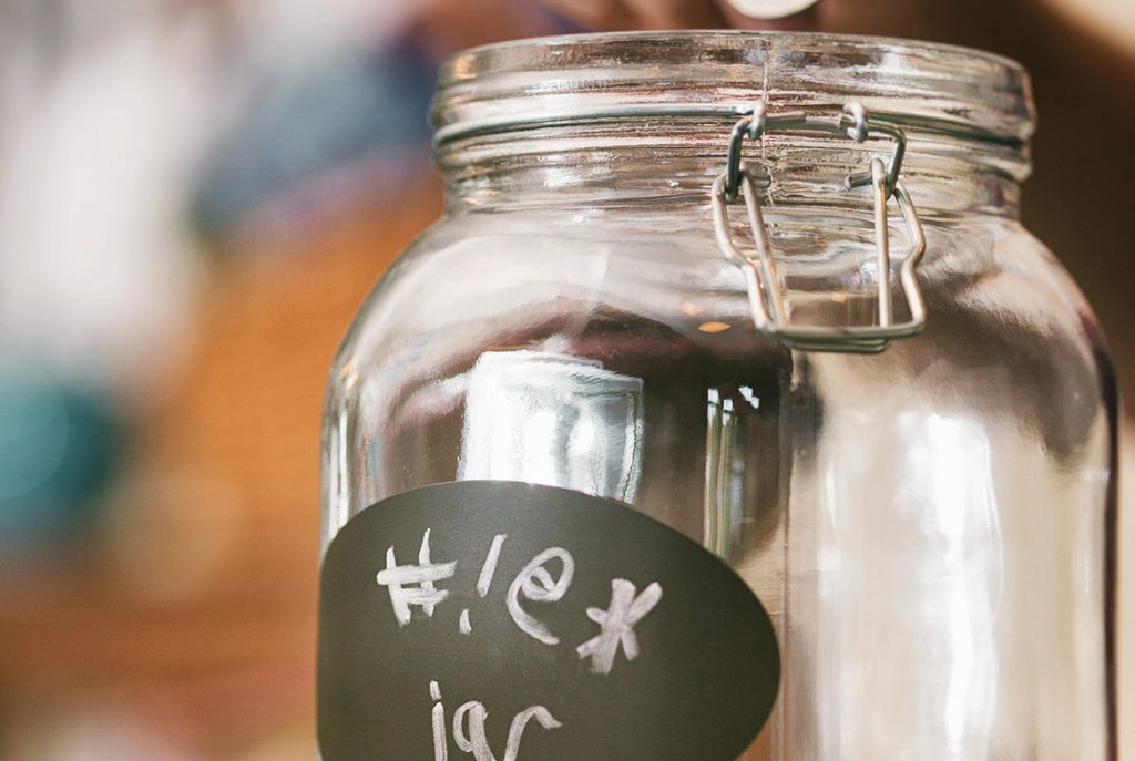 A clear glass jar with a metal latch sits on a surface. The jar has a black chalkboard label on the front with the handwritten text "#1 ex jar." The background is blurred and out of focus.