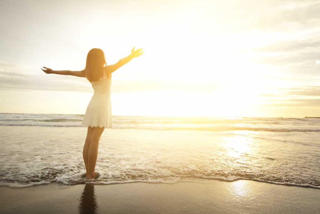 A woman in a white dress stands barefoot at the edge of the ocean, arms outstretched, facing the setting sun. The golden light reflects on the water, creating a serene and peaceful scene.