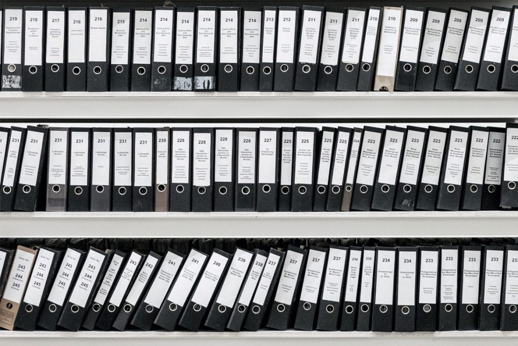 A series of white shelves filled with neatly organized black binders, each labeled with numbers and text on their spines. The binders are positioned upright, with some slightly slanted, creating a uniform and orderly appearance.