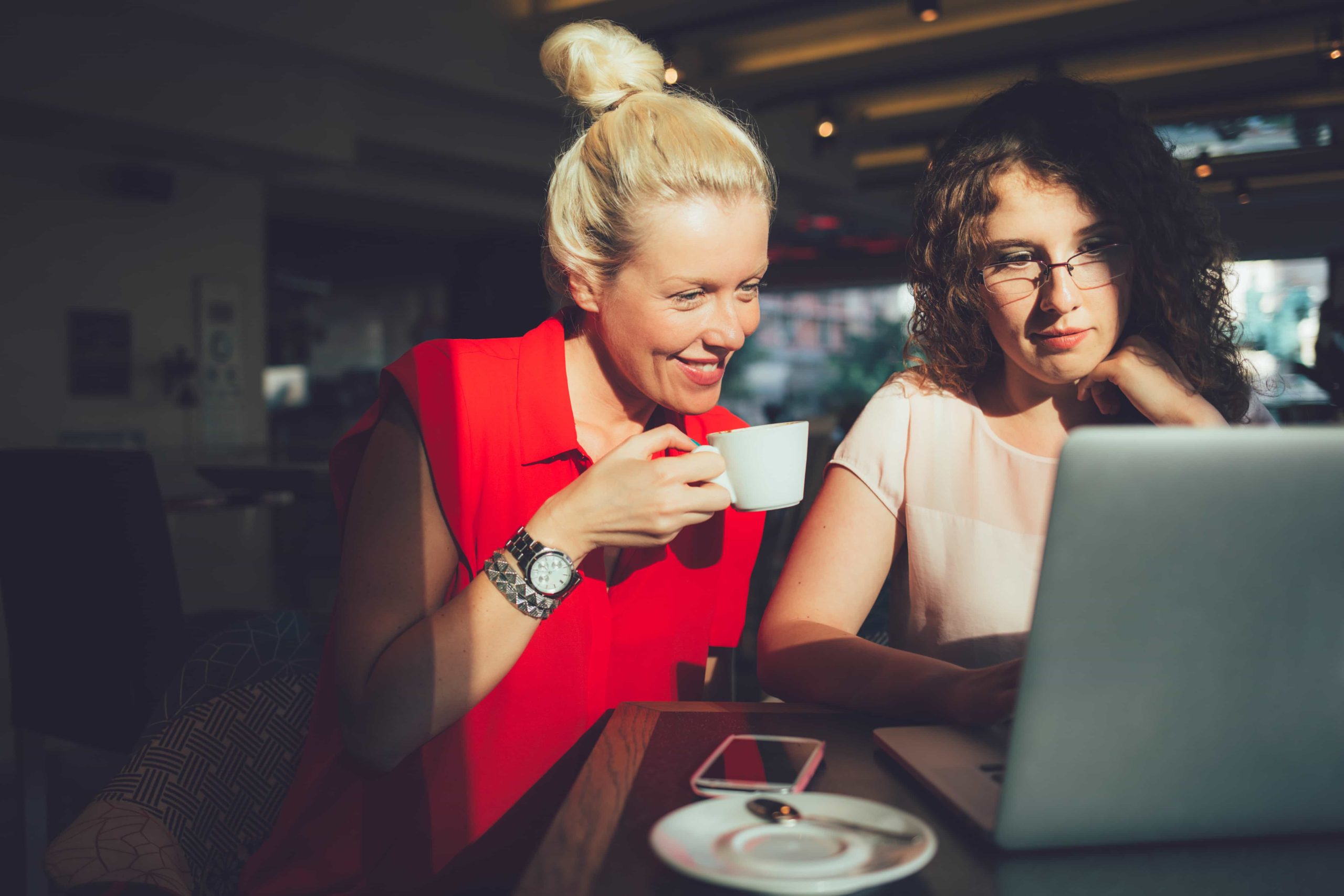 Two women are seated at a table in a café, sharing a laptop. The woman on the left, wearing a red sleeveless top, is smiling and holding a white coffee cup. The woman on the right, wearing glasses and a light pink top, looks focused on the laptop screen.