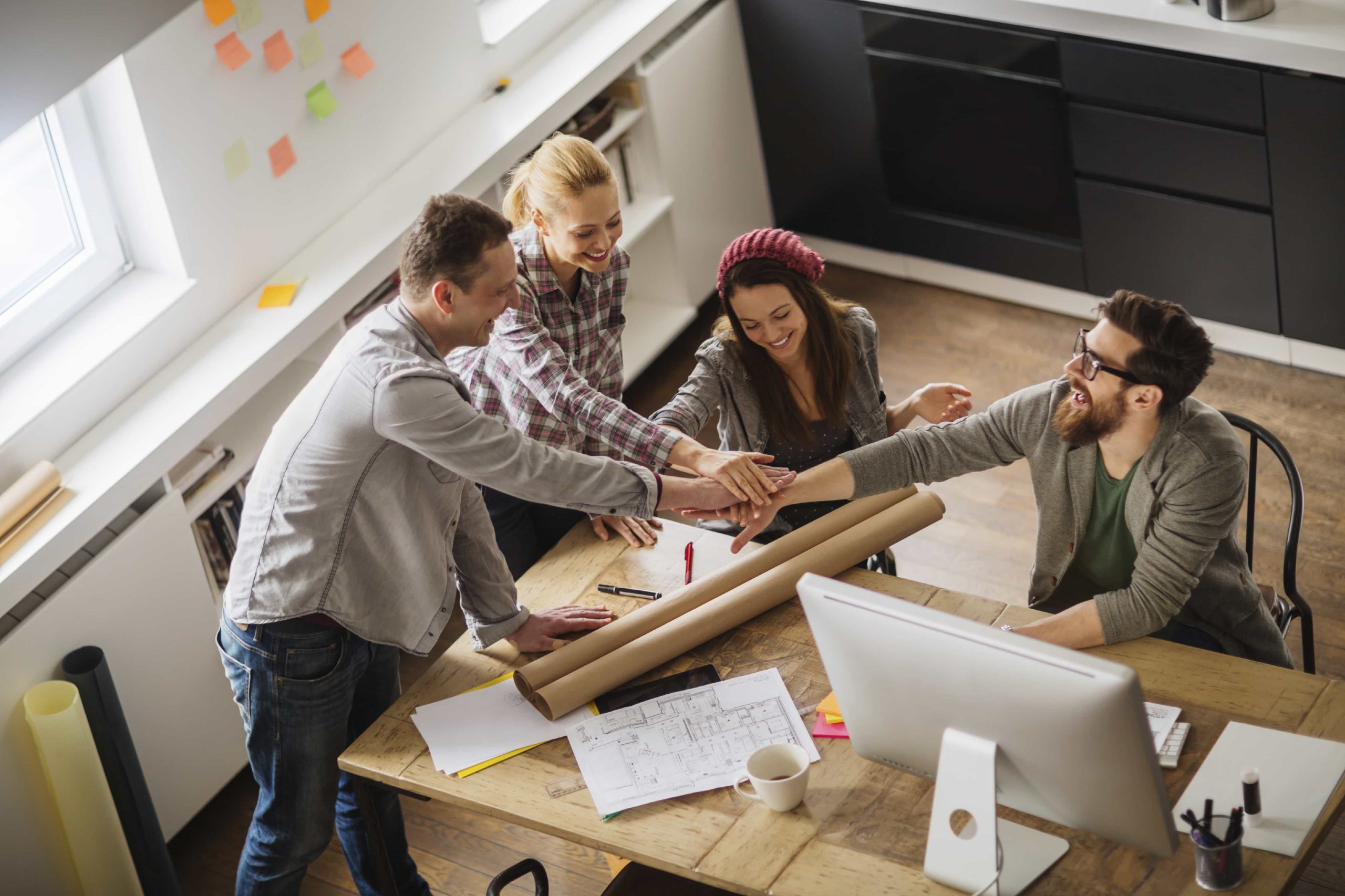 A group of four young adults in casual clothing gather around a table in a modern office. They are smiling and stacking hands together above a set of blueprints. The table also has a computer, notes, and a coffee cup. A window and sticky notes are in the background.