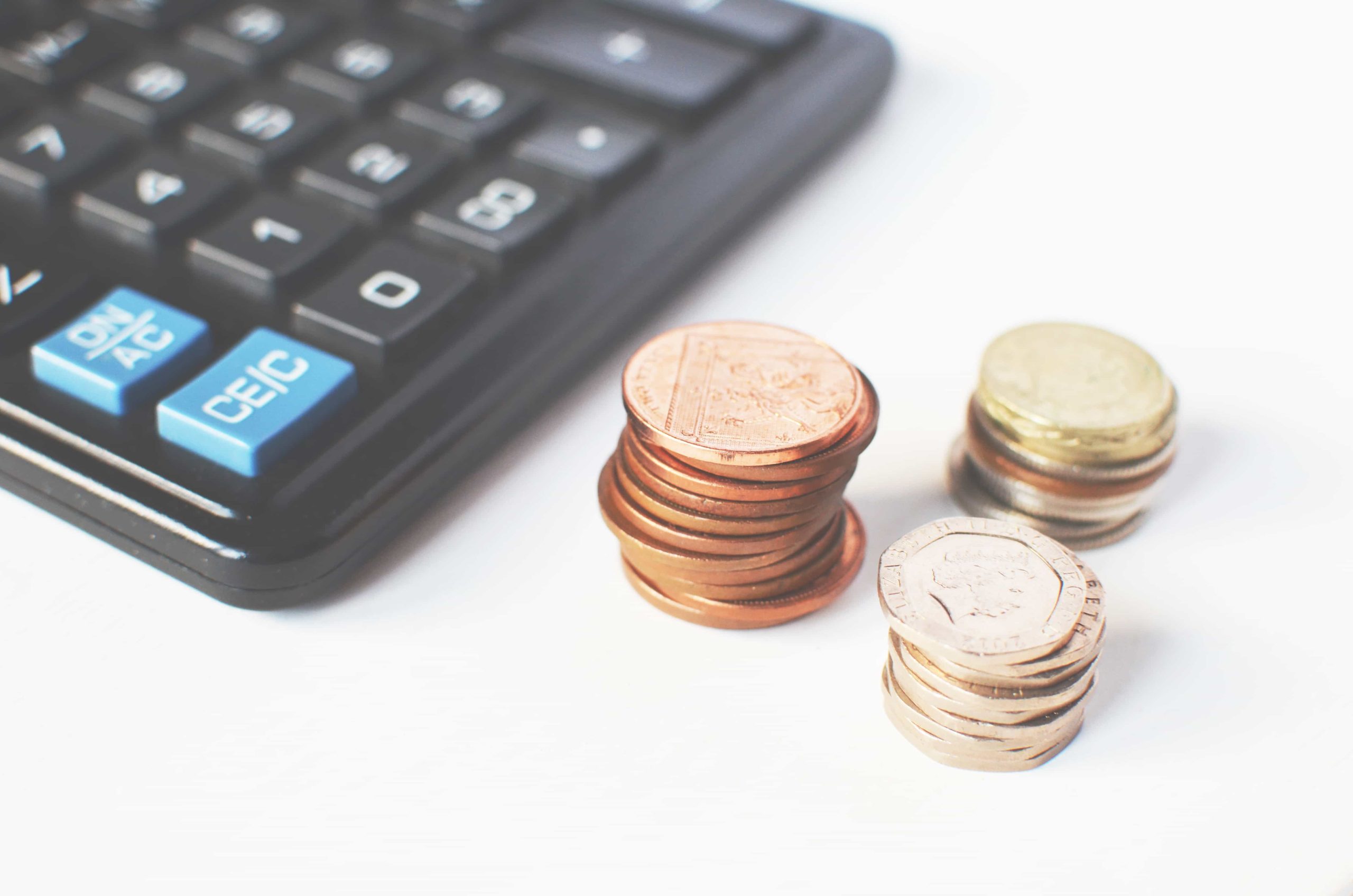 A close-up image of a calculator with a blue button labeled "CE/C" visible on the left side. On the right side, there are three small stacks of coins in copper, silver, and gold colors.