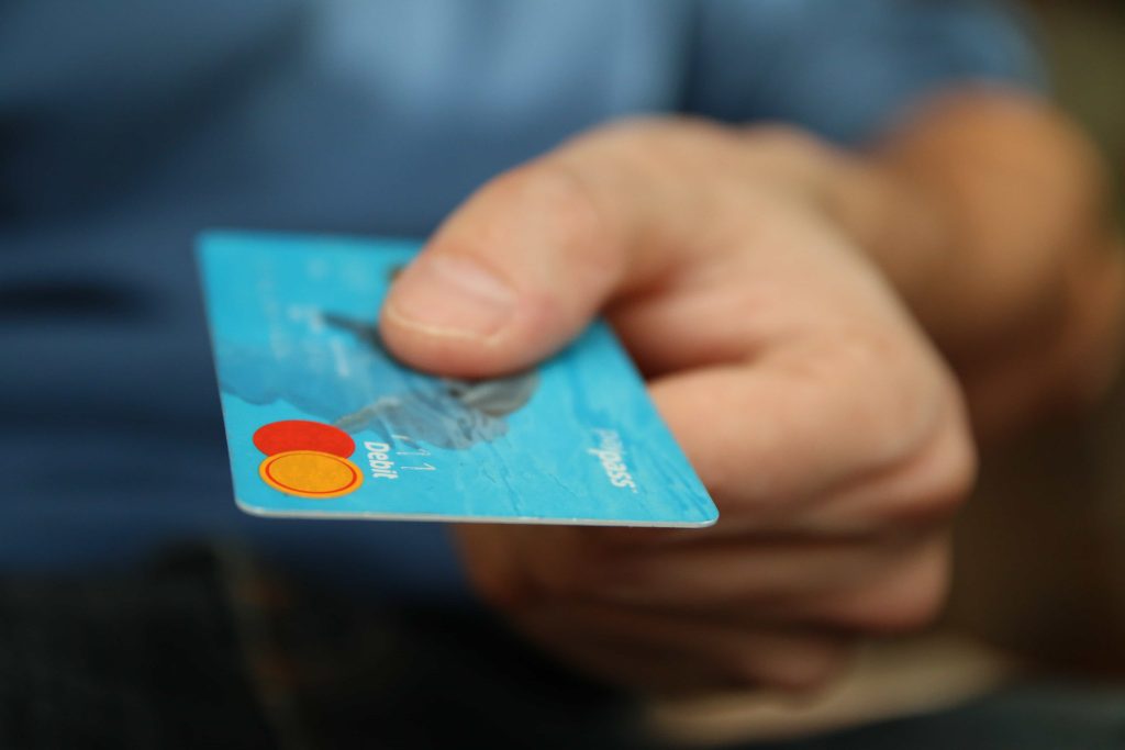 A close-up of a hand holding a blue credit card. The card is being held out towards the viewer, with visible Mastercard and Debit logos on the bottom left corner.