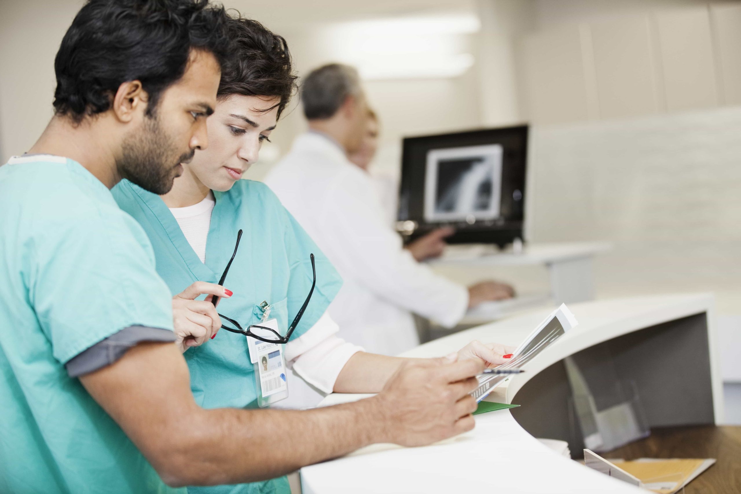 Two healthcare professionals in scrubs closely examine a document at a medical station. One holds a pair of glasses and the other holds a tablet. In the background, another medical professional is seen reviewing a scan on a monitor. Various medical equipment surrounds them.