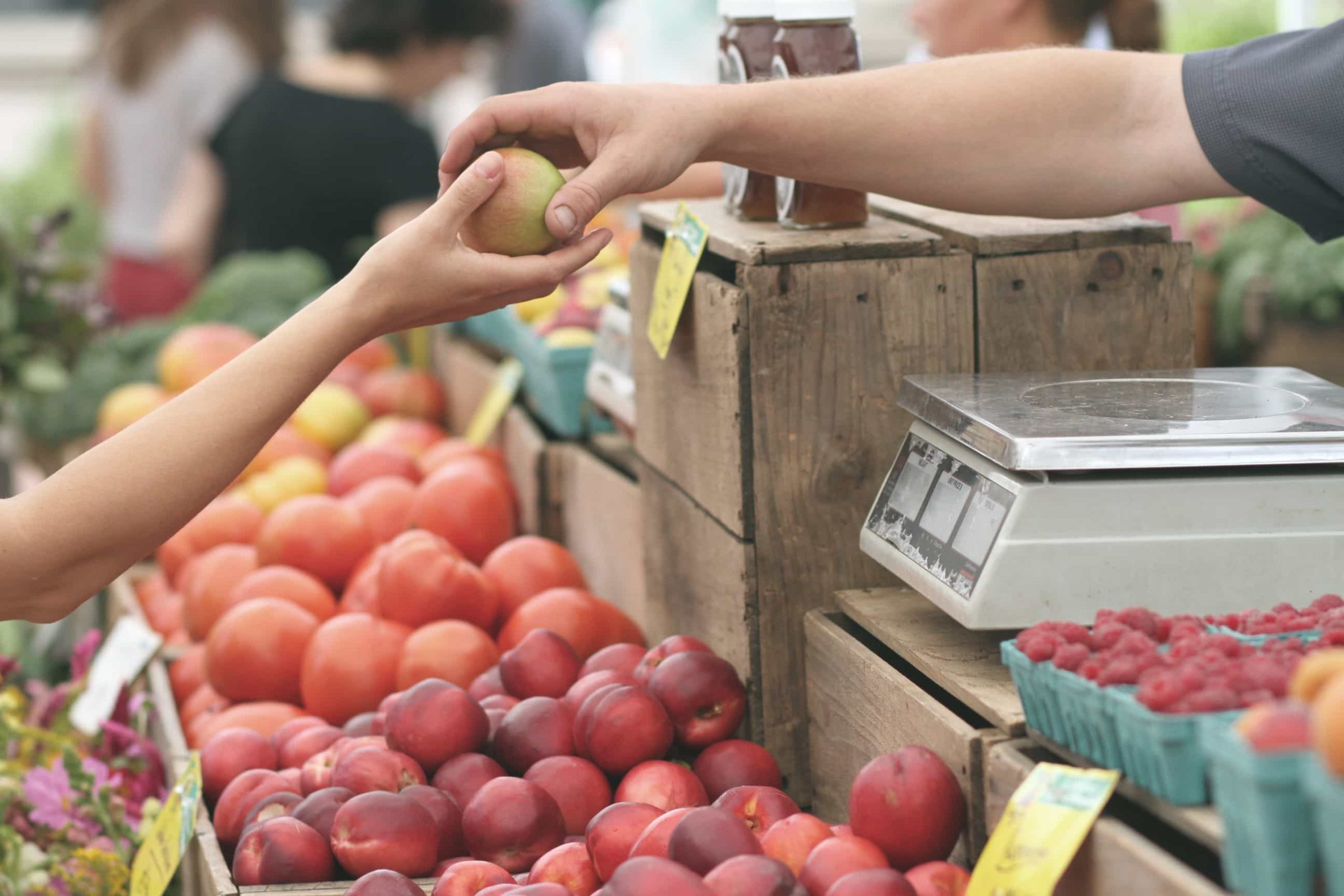 A vendor hands a piece of fruit to a customer at a stall in a farmers' market. The stall is stocked with various fresh produce, including tomatoes, peaches, and berries. A scale and price tags are visible on the wooden counter.