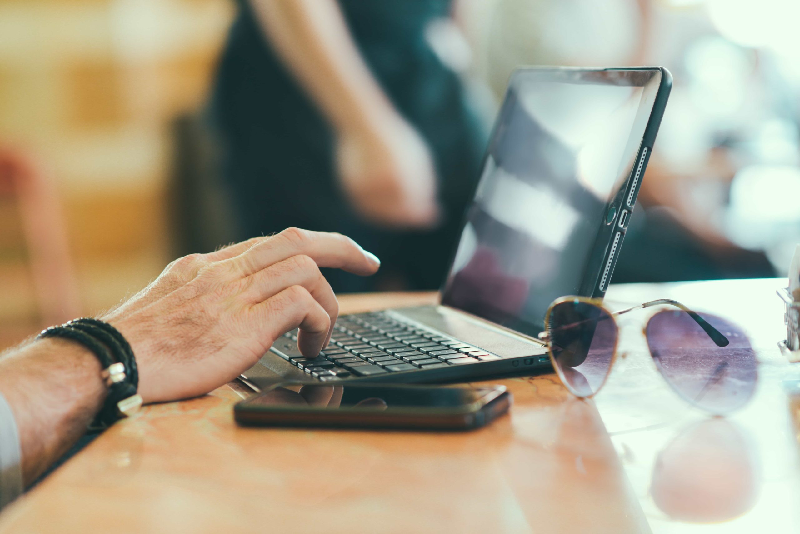 Close-up of a person's hand typing on a tablet with a detachable keyboard. The hand is adorned with bracelets. In the foreground, there are sunglasses and a smartphone on the table. The background is blurred, suggesting an indoor cafe or workspace setting.