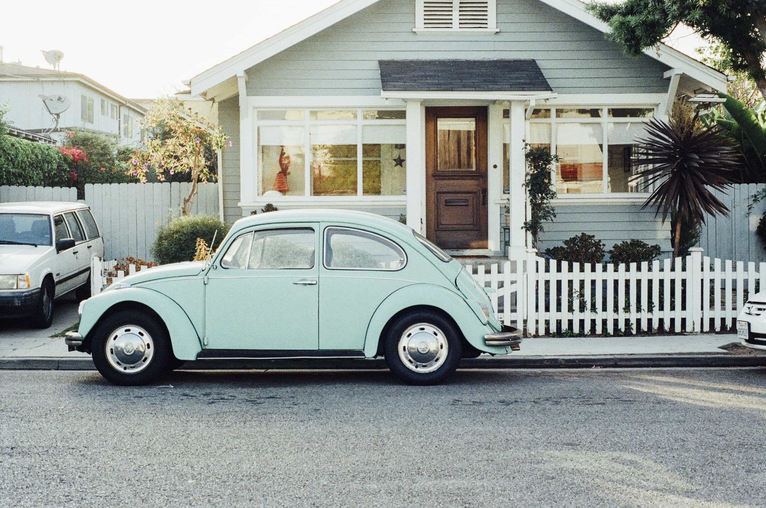 A light blue vintage Volkswagen Beetle is parked on the street in front of a charming gray house with white trim and a wooden door. The house has a white picket fence, lush greenery, and a tall tree on the right side.