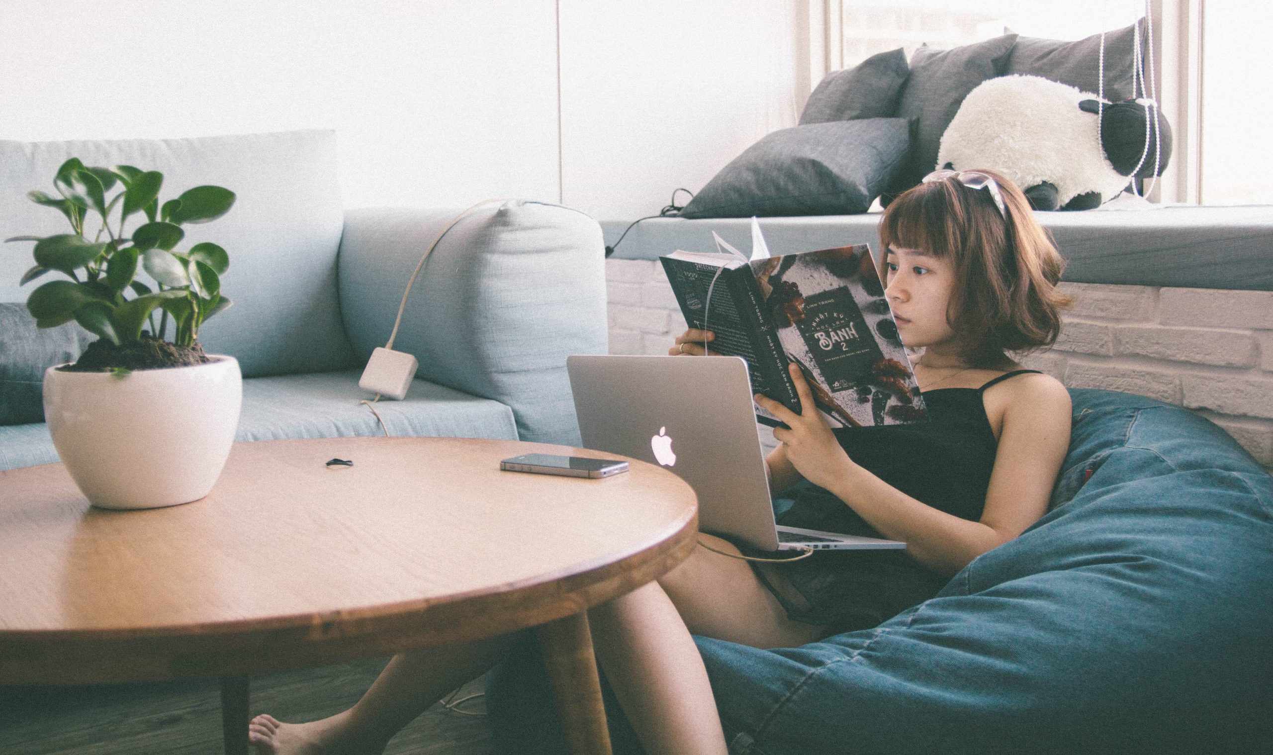 A person lounges on a bean bag chair in a cozy room, reading a book while a laptop rests on their legs. A phone lies on a wooden table in front of them next to a potted plant.