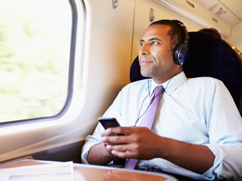 A man sitting by a train window, wearing earphones and a light blue shirt with a tie, holds a smartphone in his hands, looking out the window with a relaxed expression. Bright daylight illuminates the scene.