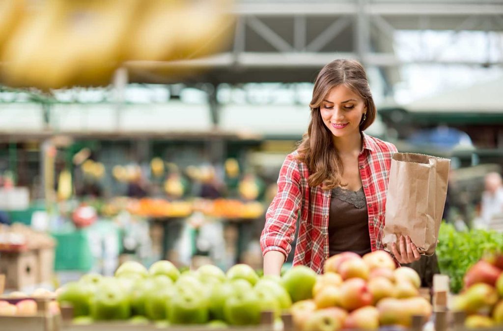 A woman in a plaid shirt is selecting apples at a farmers market. She is holding a paper bag and smiling while looking at the produce. The background shows various other fruits and market stalls.