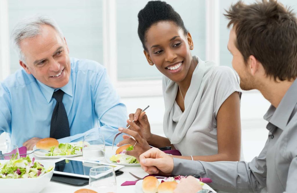 Three people are sitting at a table, smiling and talking, while having a meal. The table has plates with food, including salads and bread rolls. There is also a tablet and glasses of water on the table. The setting appears to be a professional or business environment.