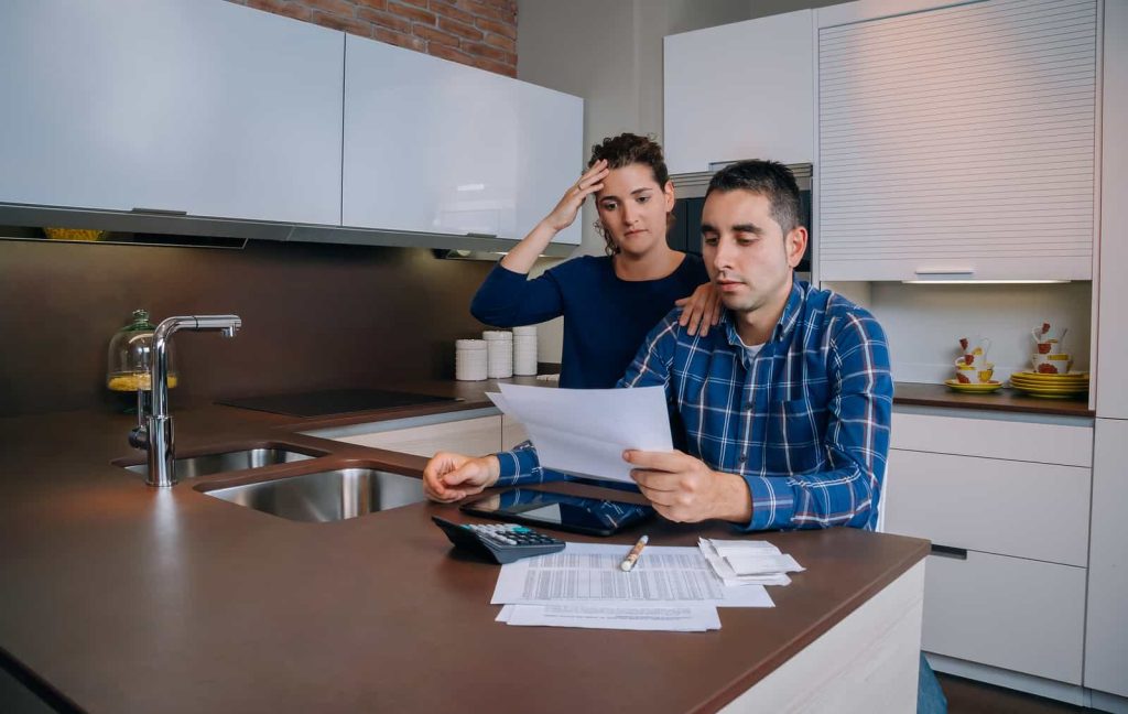 A couple stands at a kitchen counter reviewing documents. The woman, with a hand on her head, looks concerned, while the man, seated, examines the papers intently. A calculator, envelopes, and papers are spread out on the counter.