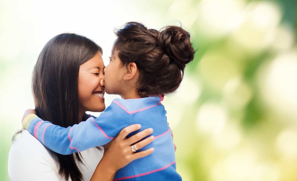 A mother and daughter share a joyful moment as they hug each other. The mother is smiling with her eyes closed, while the daughter, wearing a blue and pink striped sweater, gives her a kiss on the nose. The background is blurred greenery with bright, soft light.