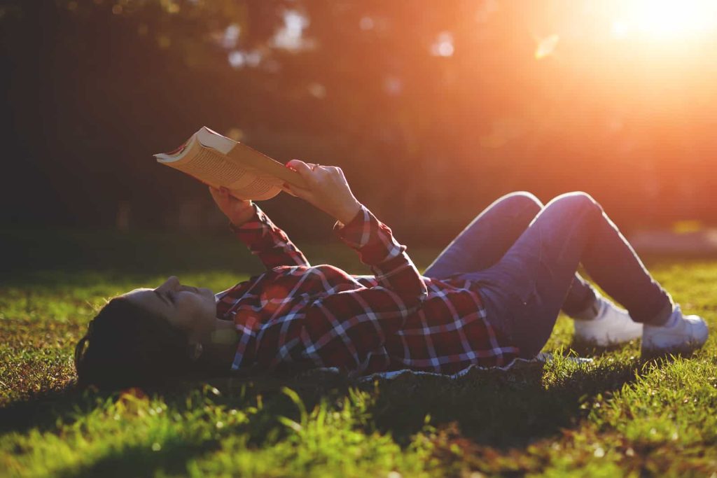 A person lying on the grass in a park, wearing a plaid shirt and jeans, reads a book held above their head. The sun is setting in the background, casting a warm, golden light over the scene.