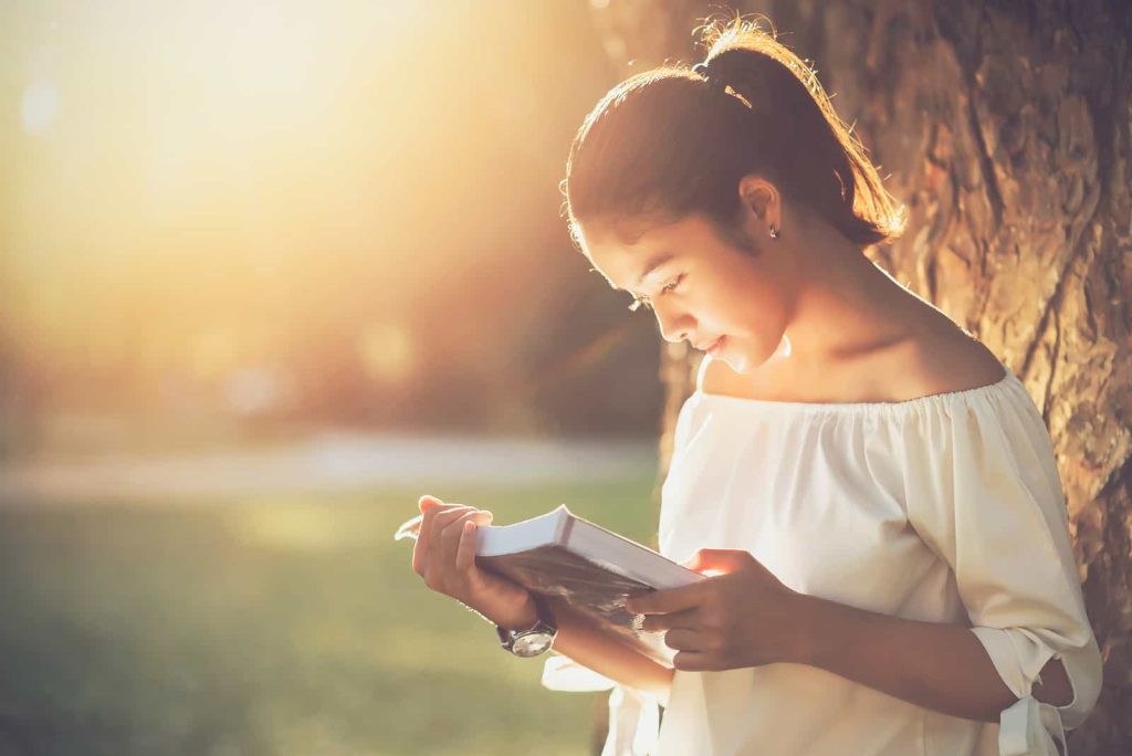 A young woman with her hair tied back is leaning against a tree while reading a book. She is wearing an off-shoulder white top and is illuminated by warm sunlight, creating a serene and peaceful atmosphere.