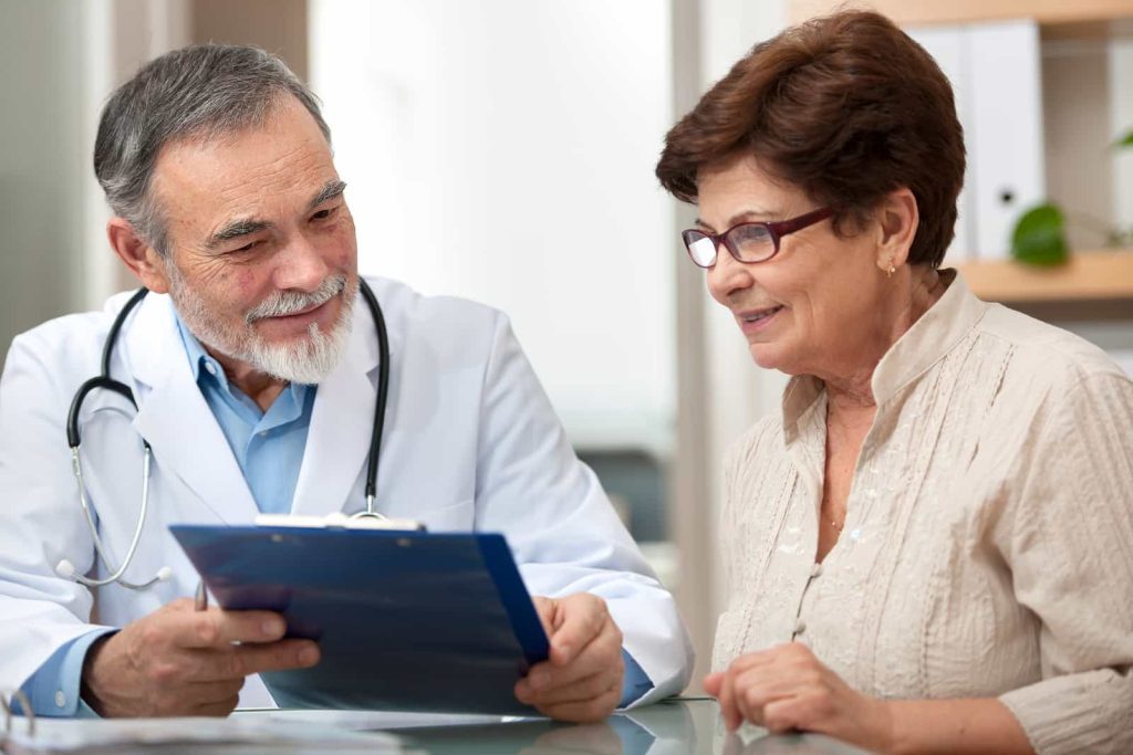 A doctor with gray hair and a white coat is holding a blue clipboard and engaging in a conversation with an elderly woman wearing glasses and a light-colored blouse. They are both seated at a desk in an office setting.