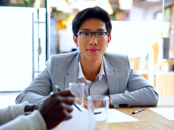 A man with glasses and short dark hair, wearing a light gray blazer and white shirt, is seated at a desk. He is looking attentively ahead. There are two people in the foreground with blurred hands, writing on paper.