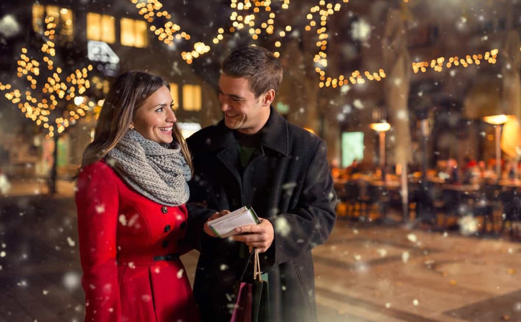 A couple stands together outdoors, smiling and holding shopping bags in a snow-covered, festive street decorated with holiday lights. The woman wears a red coat and a knit scarf, while the man wears a dark coat.