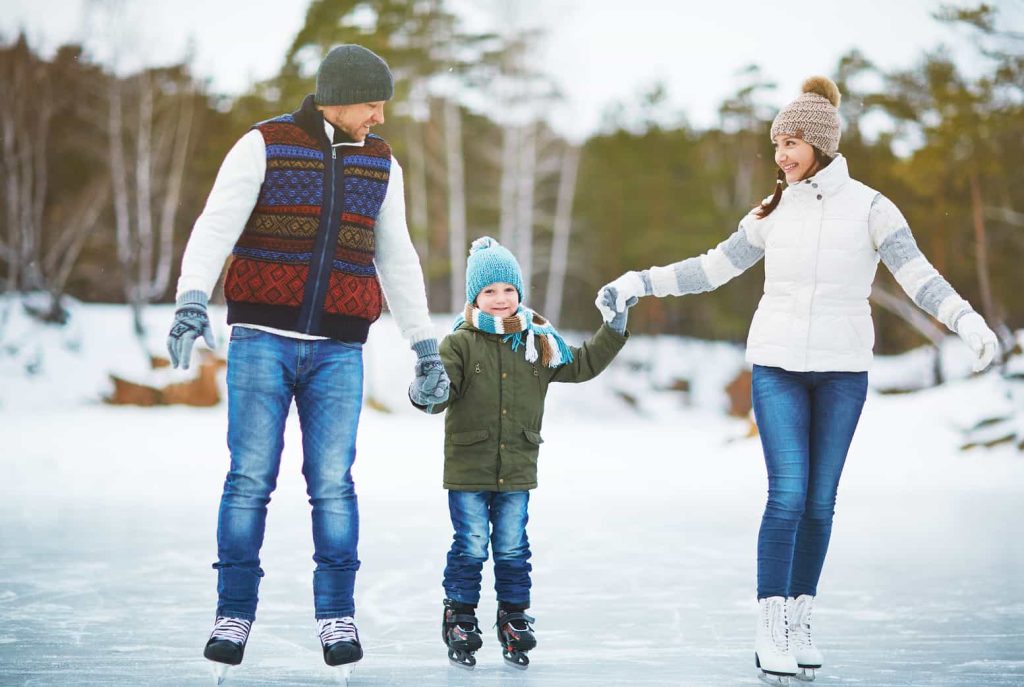 A family of three ice skating outdoors on a frozen pond in winter. A man and woman, both in warm clothing, hold hands with a young child who is wearing a blue hat, green jacket, and gloves. Snowy trees are visible in the background.