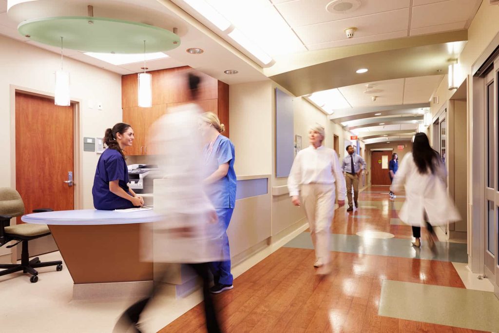 A busy hospital corridor with medical staff in scrubs and lab coats walking in various directions. A nurse sits at a reception desk, speaking with a colleague. The hallway is brightly lit and features wood and tile flooring.