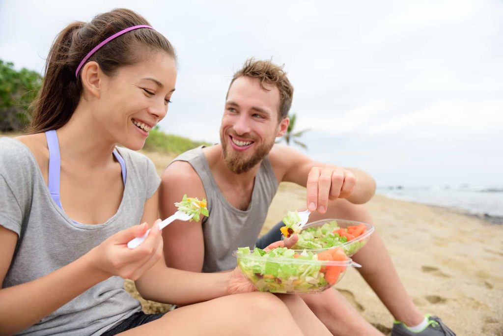 A young man and woman smiling and eating salad from a bowl on a sandy beach, with the ocean in the background. they are dressed in casual, sporty attire.