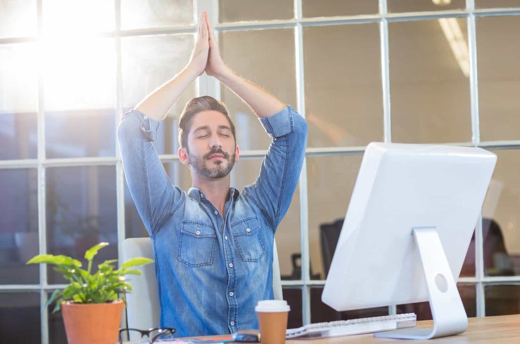 A man in a denim shirt sits at a desk in a sunlit office, eyes closed and hands clasped above his head in a stretch or relaxation pose, with a computer and coffee cup in front of him.