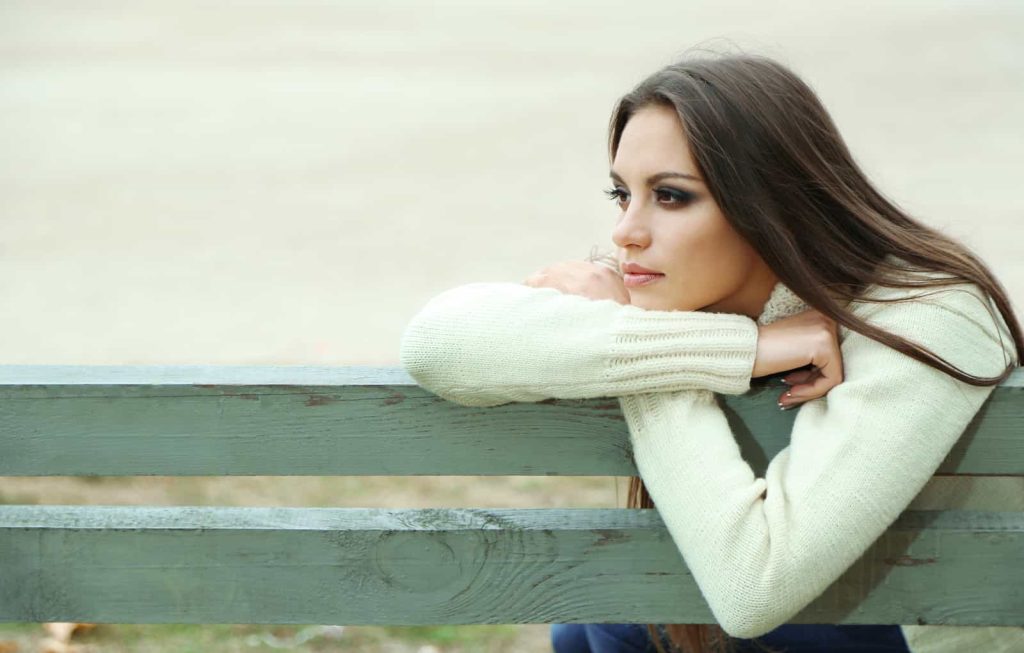 A thoughtful woman leaning on a park bench, gazing into the distance, wearing a cream colored sweater.