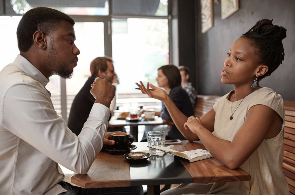 A young man and woman are engaged in a conversation at a café table, with coffee cups in front of them. The man listens intently while the woman gestures with her hand, expressing her point.