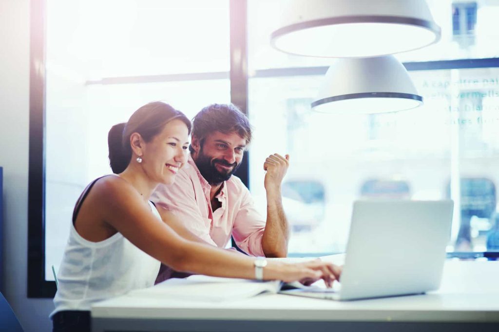 Two professionals, a woman and a man, are collaborating at a desk with a laptop in a brightly lit office space. They appear engaged and happy as they discuss their work.
