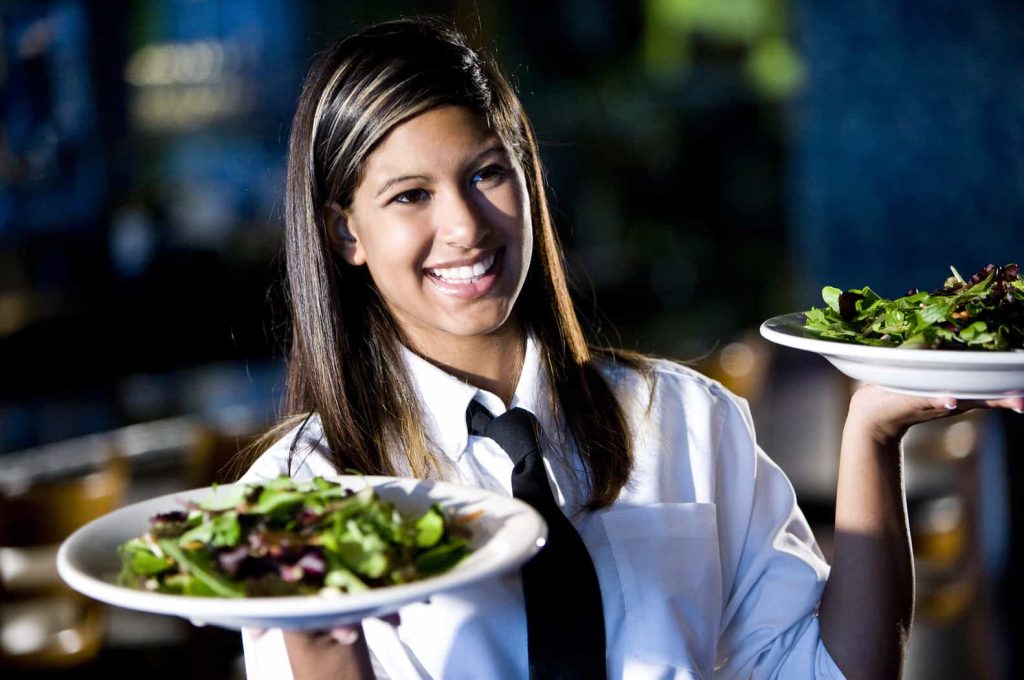 A smiling waitress in a white shirt and black tie holds two plates of salad, standing in a restaurant setting with blurred blue background.