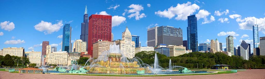 Panoramic view of a Chicago city skyline featuring modern skyscrapers, a historic building, and a large fountain in the foreground under a clear blue sky.