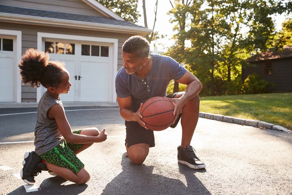 A man and a young girl playing basketball together in a driveway, with a house and garage in the background, during a sunny day.
