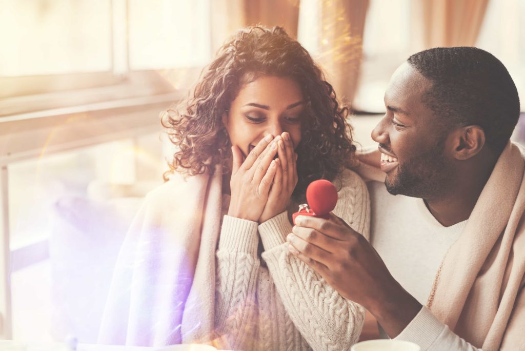 A man presents an engagement ring to a surprised woman in a cozy setting, both smiling happily. the warm sunlight filters through the window, enhancing the intimate atmosphere.