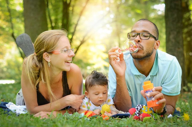 A happy family with a baby sitting on a picnic blanket in a sunny park, with the father blowing bubbles and the mother watching joyfully.