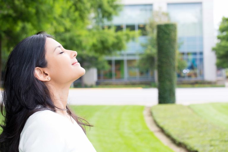 A woman with long dark hair, wearing a white blouse, closes her eyes and tilts her face upwards, enjoying the breeze outside a modern building with large windows and green lawns.