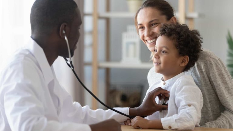 A doctor using a stethoscope on a young boy, who is smiling and sitting with his mother in a medical office.