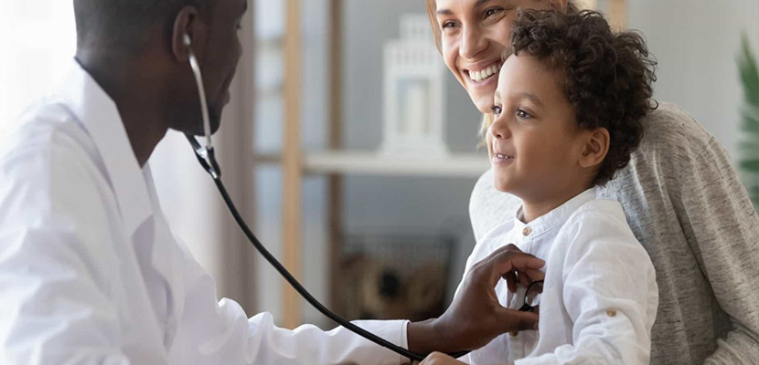 A doctor using a stethoscope on a young boy, who is smiling and sitting with his mother in a medical office.