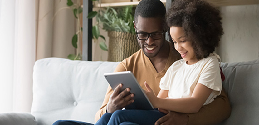 A father and a daughter, both smiling, sit on a sofa while looking at a tablet they are holding together, in a cozy room with books and plants in the background.